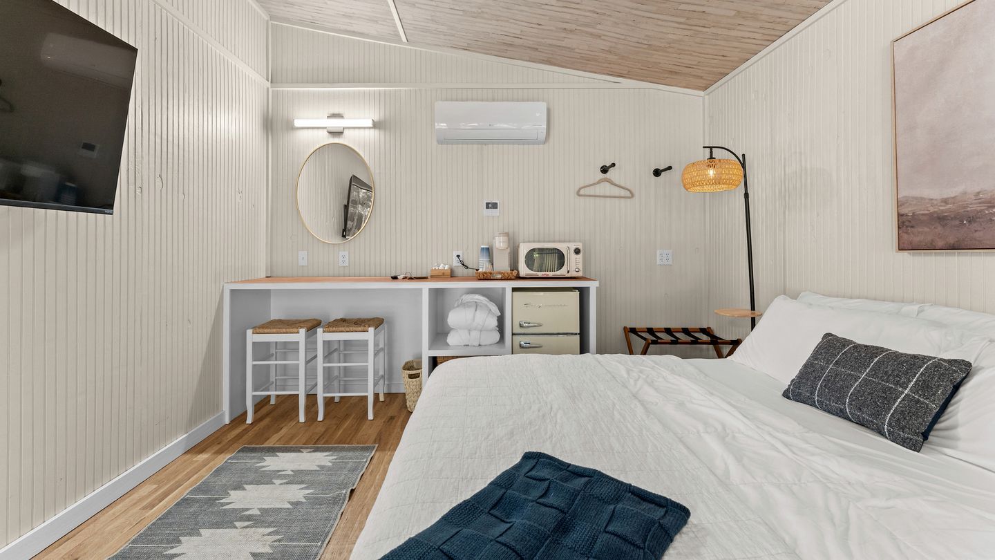 A bedroom with a bed, a desk with a chair, a hanging light fixture, and a wall-mounted air conditioner.