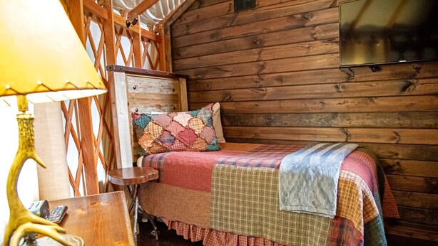 A wooden cabin room with a bed, a lamp, and a wall-mounted television. - Stunning Yurt with Magnificent Creek Views in Topton, North Carolina (Topton)