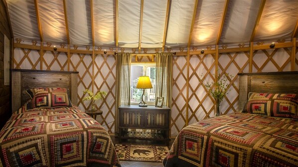 A yurt with two beds, a wooden bench, a window with curtains, and a lamp.