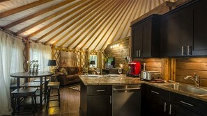 Ein gemütliches Interieur mit Holzbalkendecke, einer Essecke mit rundem Tisch und Stühlen, einer Küche mit dunklen Schränken und einem roten Gerät sowie einer Holzwand.