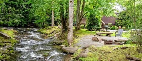A river flowing through a forest with a picnic area and a building in the background.