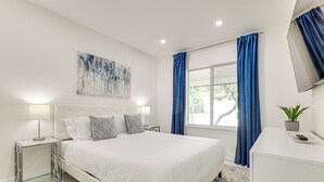 A modern bedroom with a large bed, a bedside table with a lamp, a TV on the wall, and a window with blue curtains. - 1 Mi to Las Olas Eats! Modern Apt w/ Shared Patio (Fort Lauderdale)