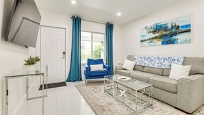 A modern living room with a glass coffee table, a blue armchair, a grey sofa, and a large abstract painting on the wall. - 1 Mi to Las Olas Eats! Modern Apt w/ Shared Patio (Fort Lauderdale)
