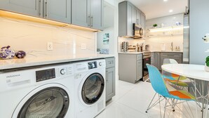 A modern kitchen with a washing machine, grey cabinets, and a dining area with colorful chairs. - 1 Mi to Las Olas Eats! Modern Apt w/ Shared Patio (Fort Lauderdale)