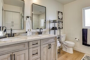 A modern bathroom with a large mirror, two sinks, a toilet, and a shelf with toiletries. - Sports Court + Hot Tub: Luxe Scottsdale Retreat! (Scottsdale)