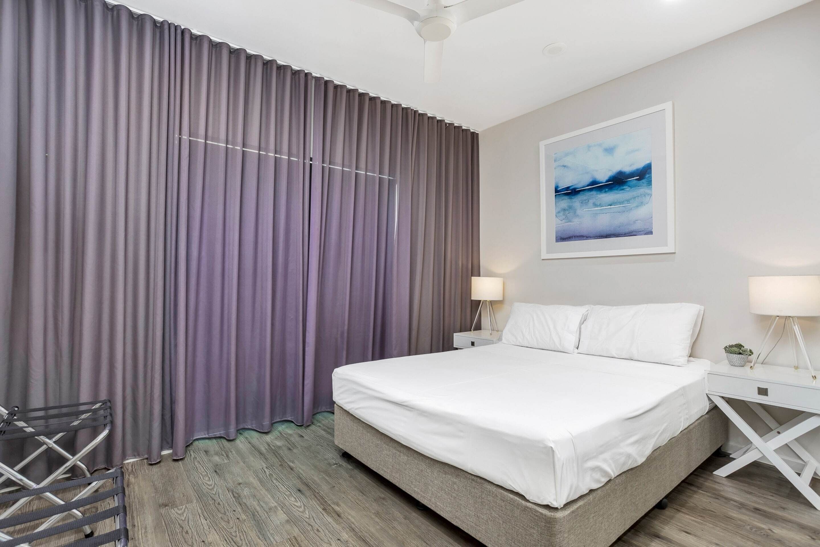 A modern hotel room with a large bed, two bedside tables with lamps, a ceiling fan, and a large window with purple curtains.
