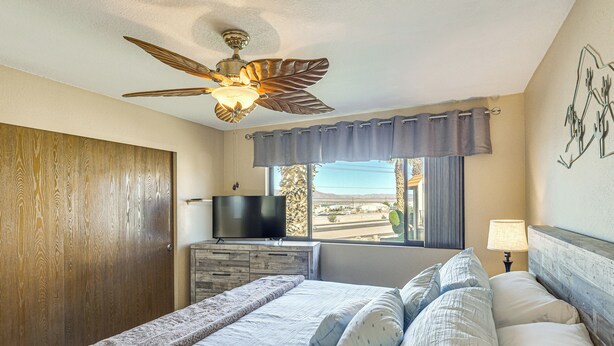 A bedroom with a bed, a ceiling fan, a TV, a window with curtains, and a wall-mounted lamp. - Pool, Lake & Sun! Havasu Hot Spot By London Bridge (Lake Havasu City)