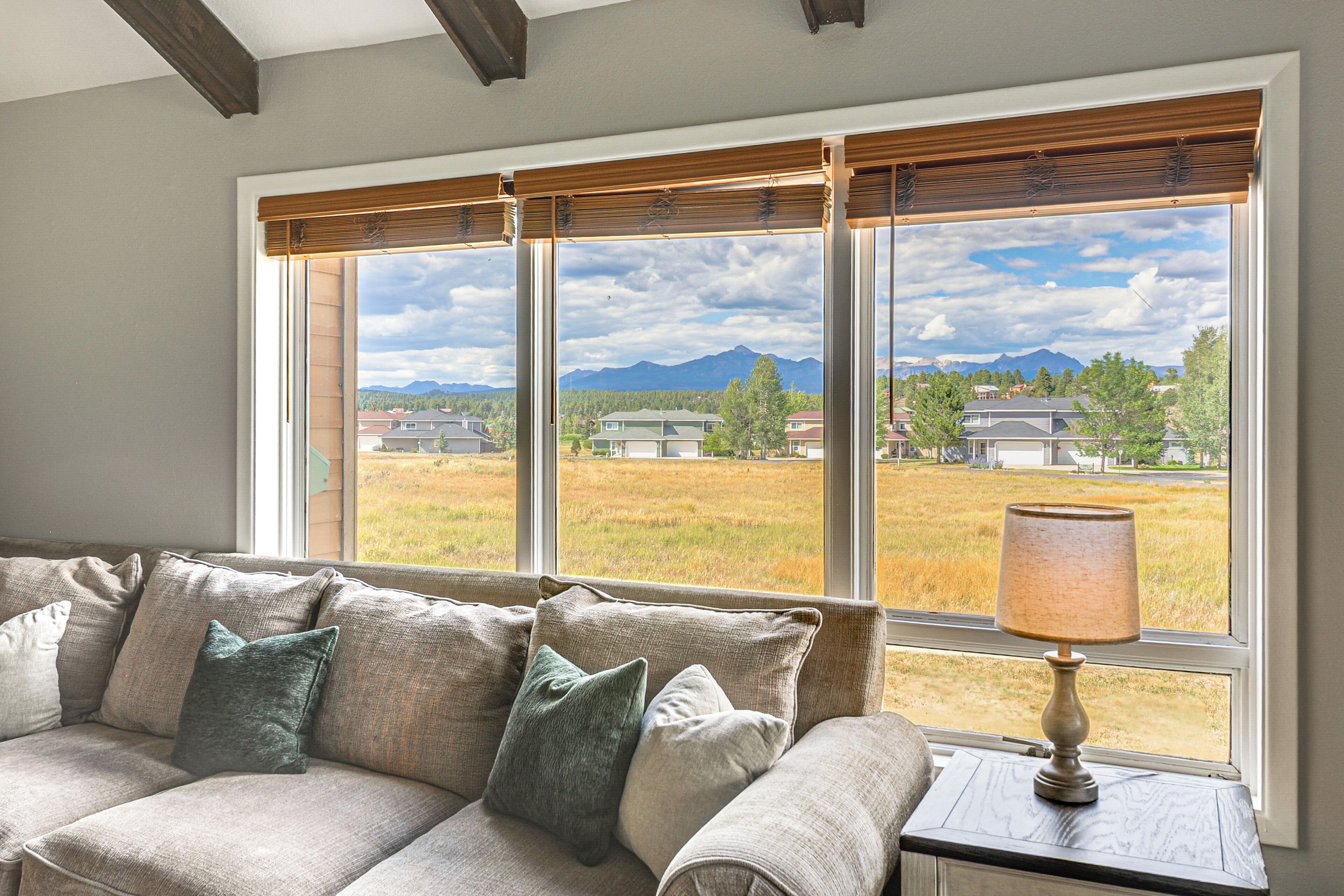 A room with a sofa facing a large window with a view of a residential area and mountains.