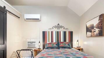 A bedroom with a bed, a headboard, a wall-mounted air conditioner, a bedside table with a lamp, and a painting on the wall.
