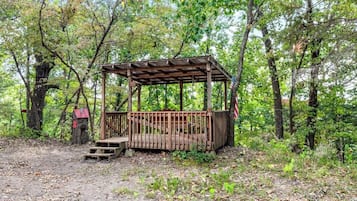 A wooden gazebo in a forested area with a dirt ground and trees in the background.