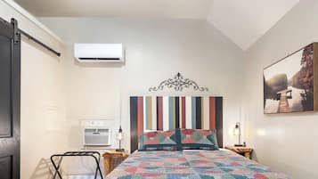 Een slaapkamer met een bed, hoofdbord, een wandairconditioner, nachtkastje met lamp en een schilderij aan de muur.
