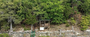 A wooden cabin with a green roof, surrounded by dense forest, and a stone wall at the base.