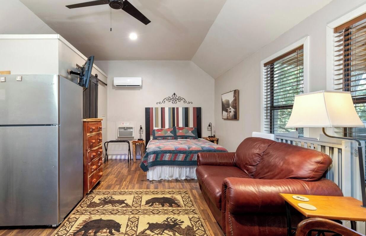 A compact room with a bed, a leather sofa, a small table, a refrigerator, a ceiling fan, and a window with blinds.