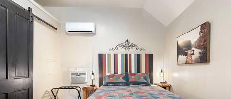 Une chambre à coucher comprenant un lit, une tête de lit, un appareil de climatisation, une table de chevet avec une lampe et un tableau accroché au mur.