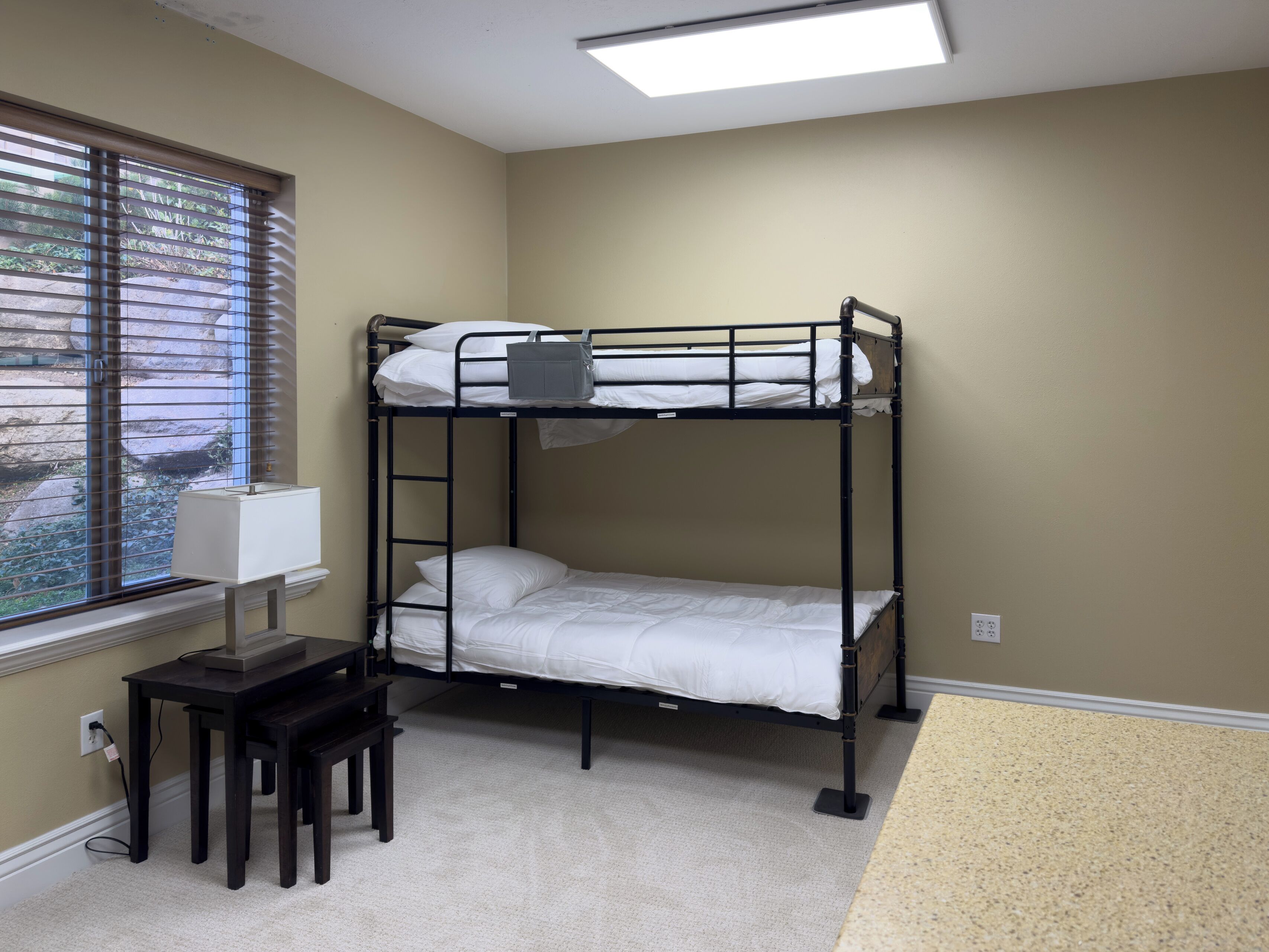 Une chambre avec un lit superposé, un bureau et une chaise, une fenêtre avec des stores et un luminaire au plafond.