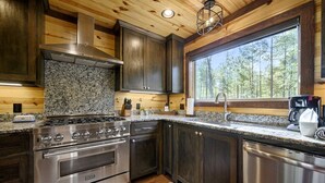 A modern kitchen with granite countertops, stainless steel appliances, and a large window offering a view of trees. - Creekside Cabin Perfect for Family Getaways in Broken Bow, Oklahoma (Broken Bow)