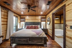 Une chambre dans un chalet en bois, comprenant un lit, des tables de chevet, un ventilateur de plafond, une fenêtre et une salle de bain avec baignoire.