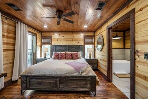 Ein Schlafzimmer einer Holzhütte mit einem Bett, Nachttischen, einem Deckenventilator, einem Fenster und einem Badezimmer mit einer Badewanne.