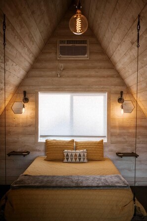 A bedroom with a sloped ceiling, a large window, a bed with multiple pillows, and wall-mounted lighting. - Cute A-Frame Cabin for an Amazing Couples Getaway near Yellowstone National Park in Emigrant, Montana (Emigrant)