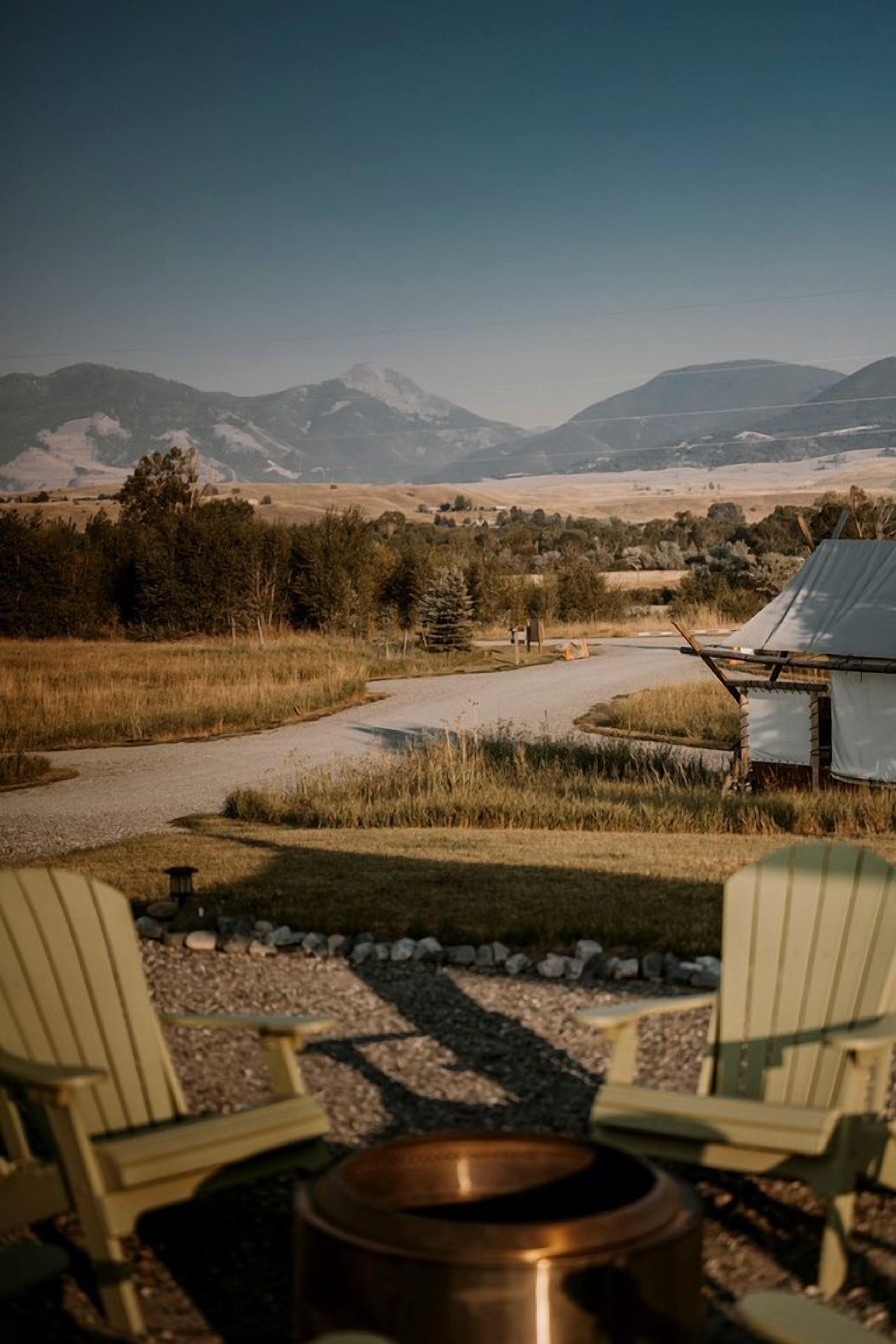 A rustic outdoor setting with a gravel path, a metal fire pit, and two Adirondack chairs facing a mountainous landscape.