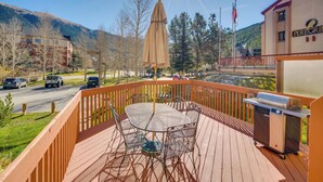 A wooden deck with a grill, outdoor table, and chairs, overlooking a road and mountains. - Ski Copper Mountain! Alpine-chic Condo w/ Hot Tub (Copper Mountain)