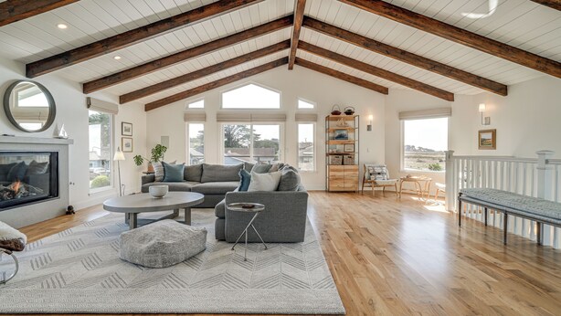 Ein geräumiges Wohnzimmer mit hohen Decken, Holzbalken, einem Kamin, einem großen Sofa, einem Couchtisch und Blick ins Freie.