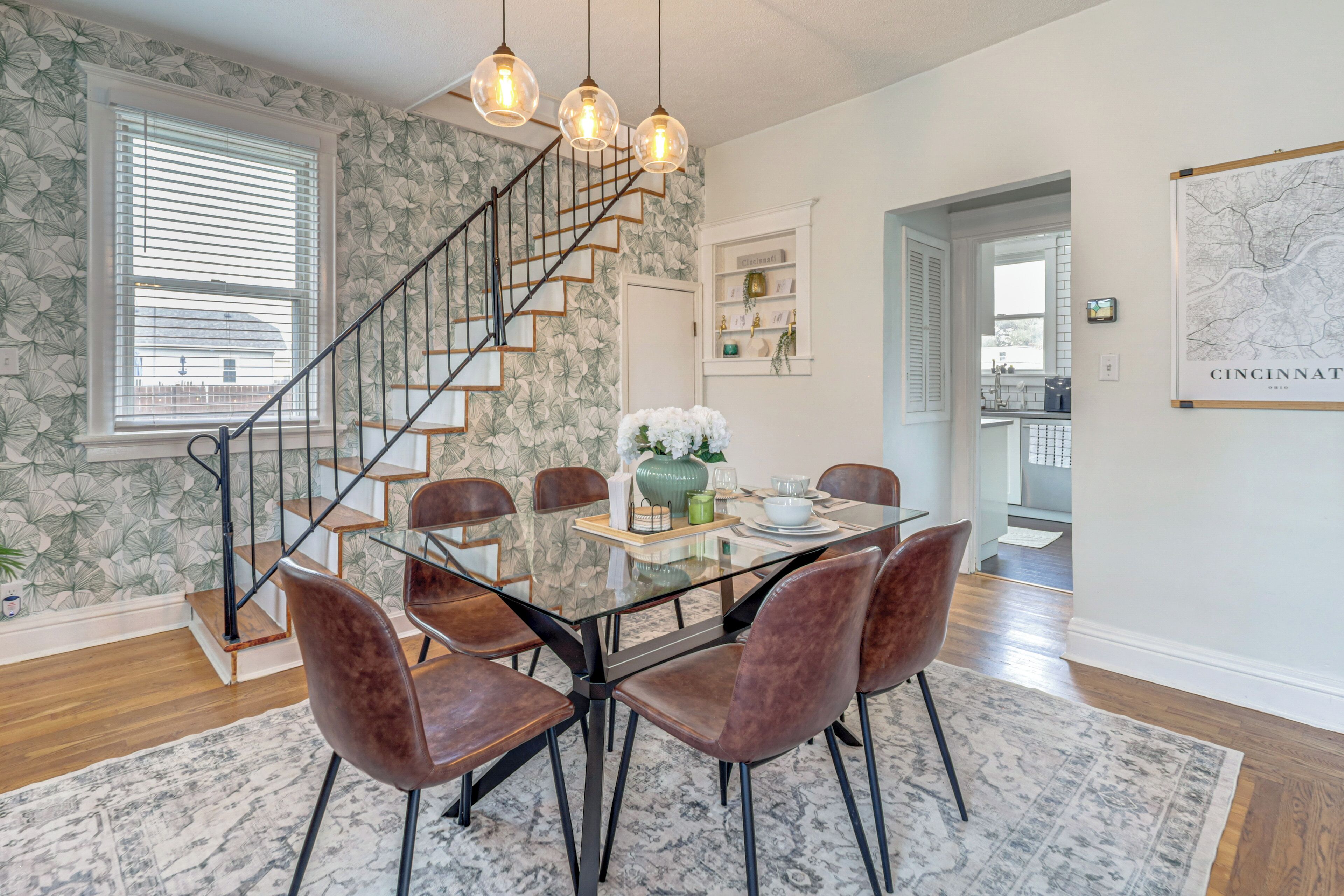 Un comedor con mesa de vidrio, sillas de cuero, una escalera y una pared con estampado floral.