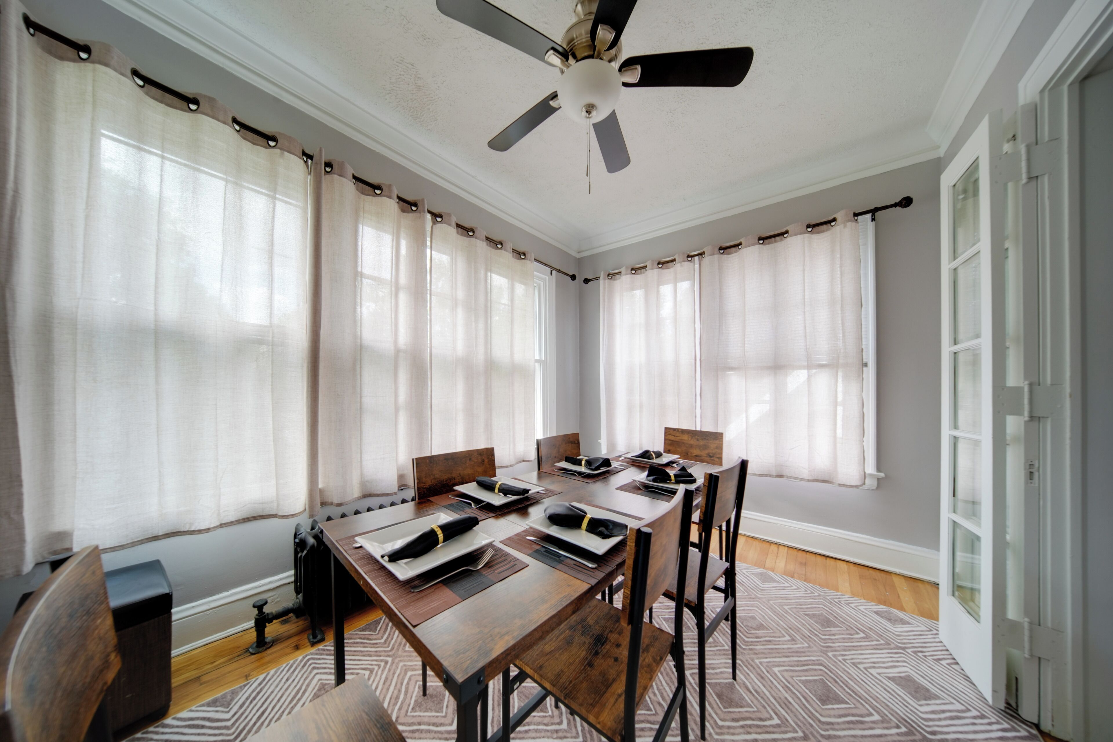 A conference room with a wooden table, chairs, a ceiling fan, and large windows with sheer curtains.