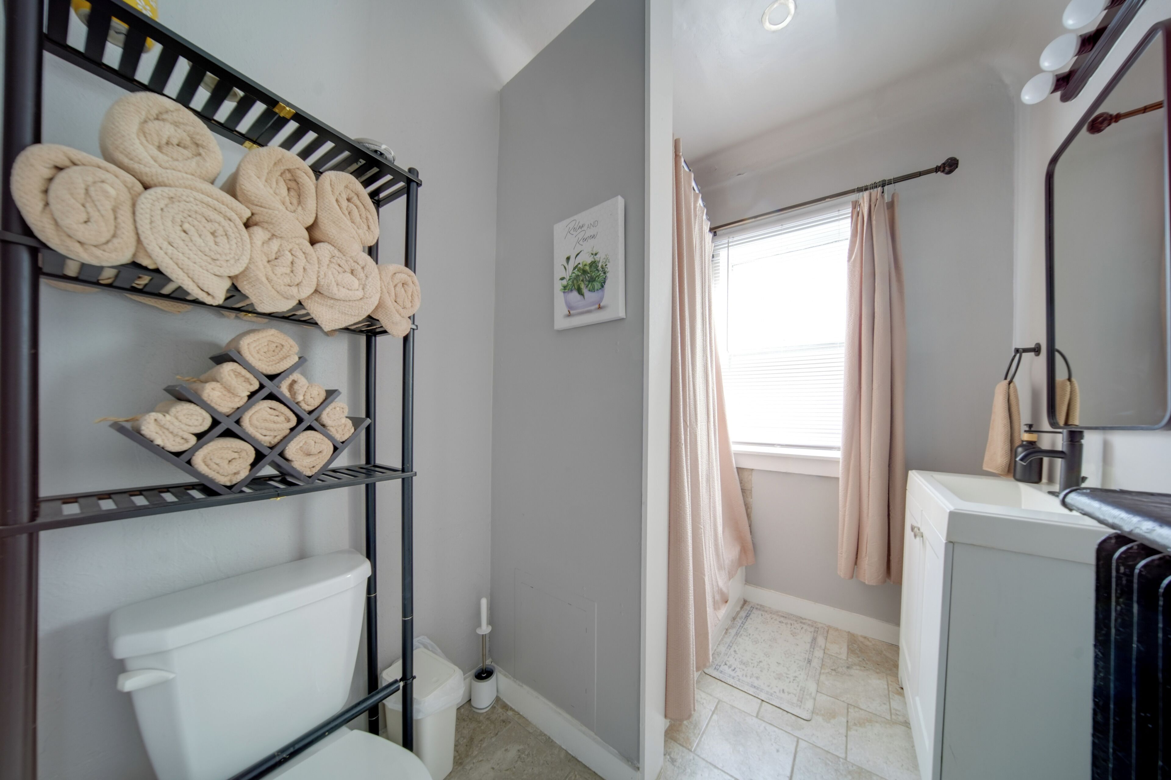 Une salle de bain comprenant des toilettes, un lavabo, un miroir, un porte-serviettes avec des serviettes, une brosse pour les toilettes et une fenêtre avec des rideaux.