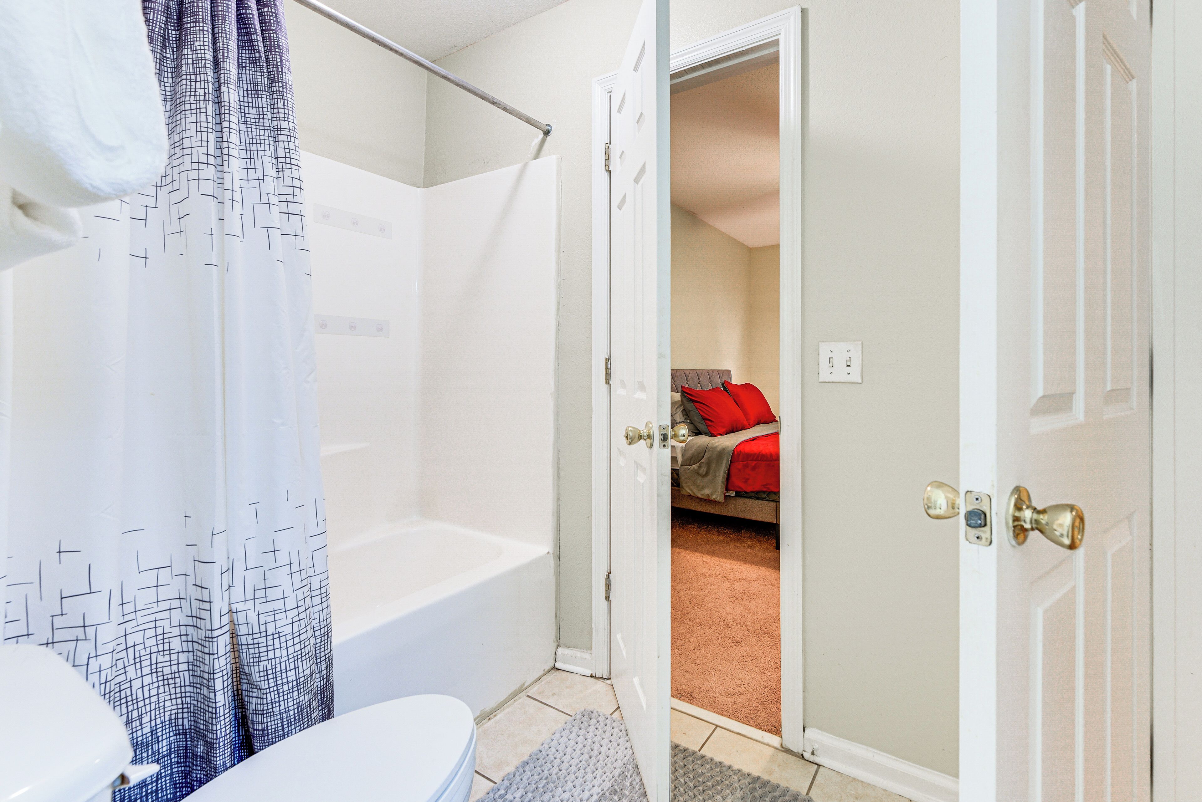 A bathroom with a shower, toilet, and a door leading to a bedroom with a bed and a red pillow.