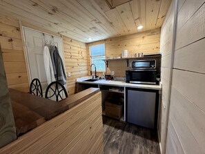 A compact kitchen with wooden walls, a microwave, a sink, and a small refrigerator. - Cozy Tiny Cabin with Rustic Charm near Mineral Wells, Texas (Mineral Wells)