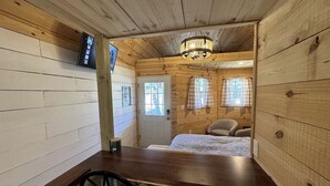 L’intérieur d’un chalet en bois comprend une table à manger, deux chaises, un lit recouvert d’une couverture à carreaux, un lustre et une porte avec des fenêtres.