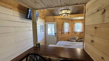 Ein Holzhütten-Interieur mit Esstisch, zwei Stühlen, einem Bett mit kariertem Überwurf, einer Pendelleuchte und einer Tür mit Fenstern.