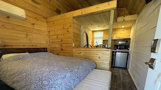 L’intérieur d’une cabane en bois, avec un lit, un banc, un coin cuisine et une porte.