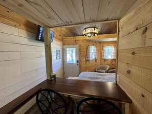 L’intérieur d’un chalet en bois comprend une table à manger, deux chaises, un lit recouvert d’une couverture à carreaux, un lustre et une porte avec des fenêtres.