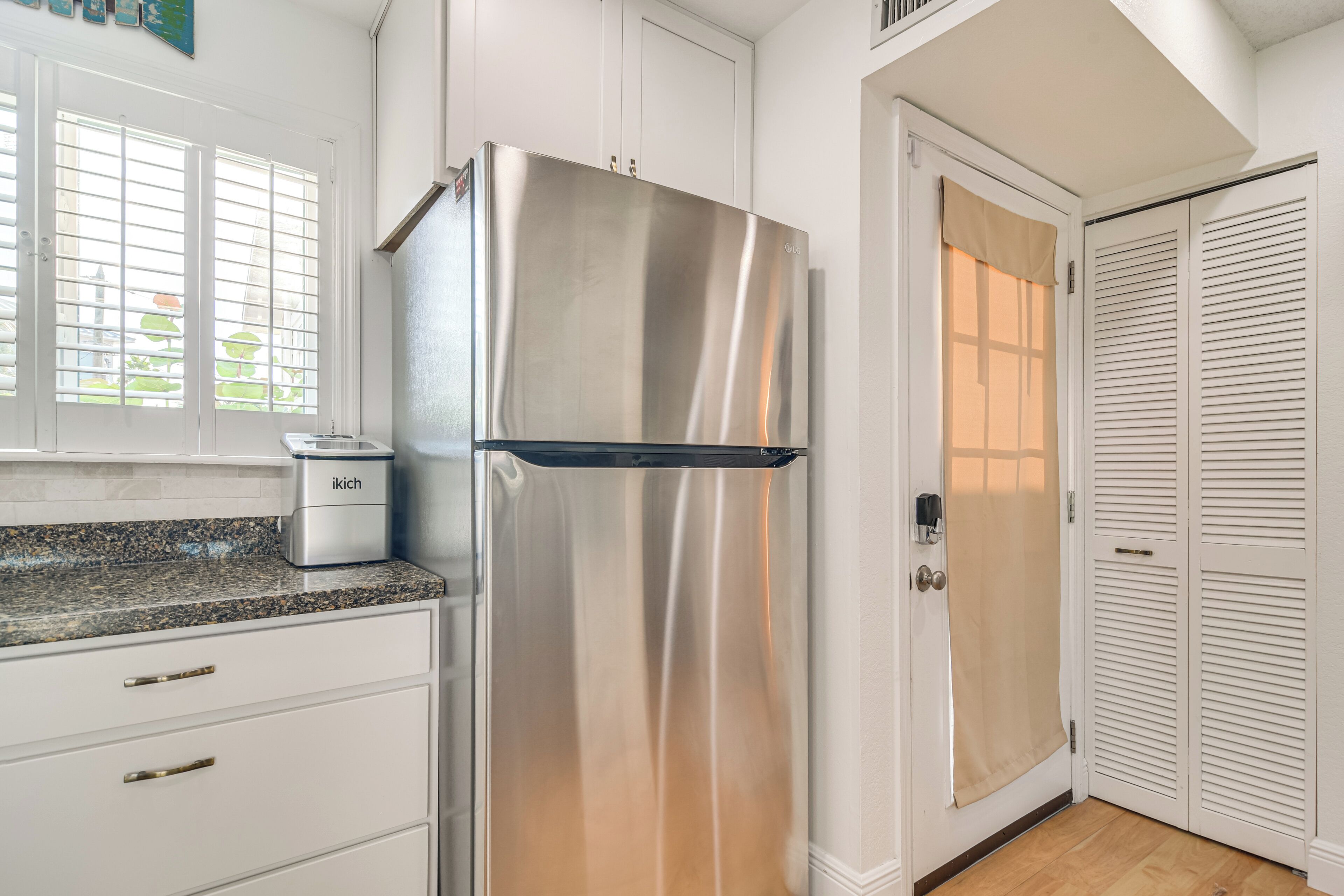 A kitchen with white cabinets, a stainless steel refrigerator, a built-in oven, and a window with blinds.