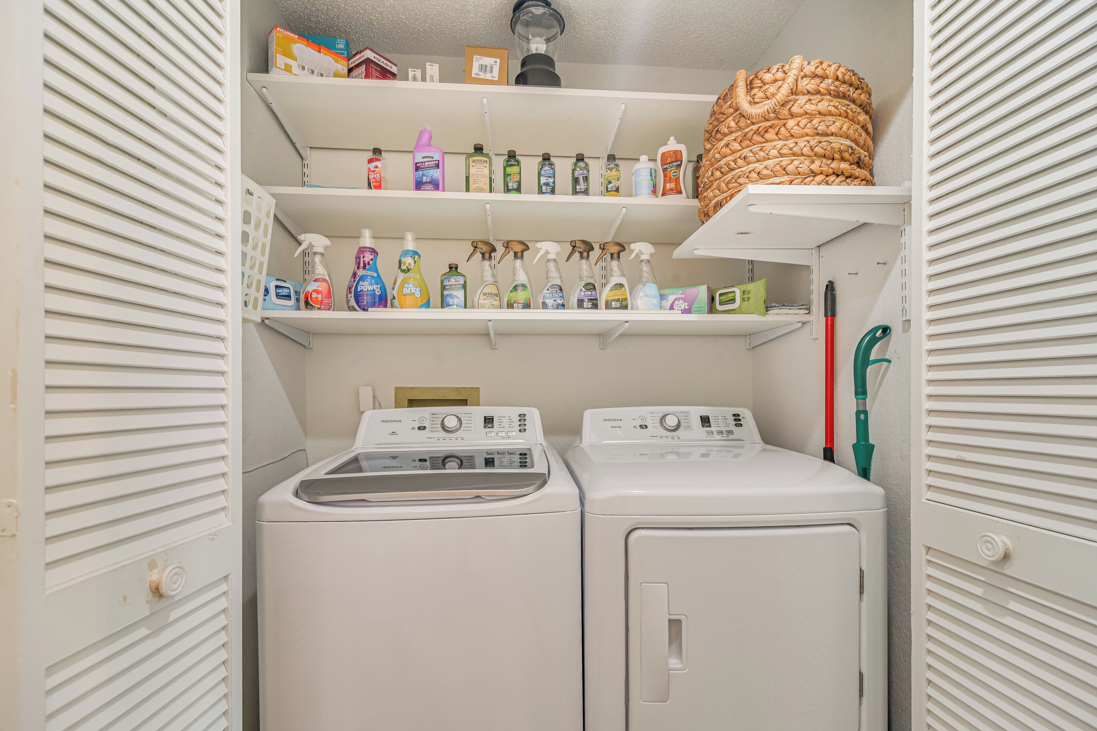 A laundry room with two washing machines, shelves with cleaning supplies, and a basket on the top shelf.