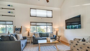 Una sala de estar con sofá, mesa de centro, sillón estampado, ventanas amplias y un televisor de pantalla plana en la pared.