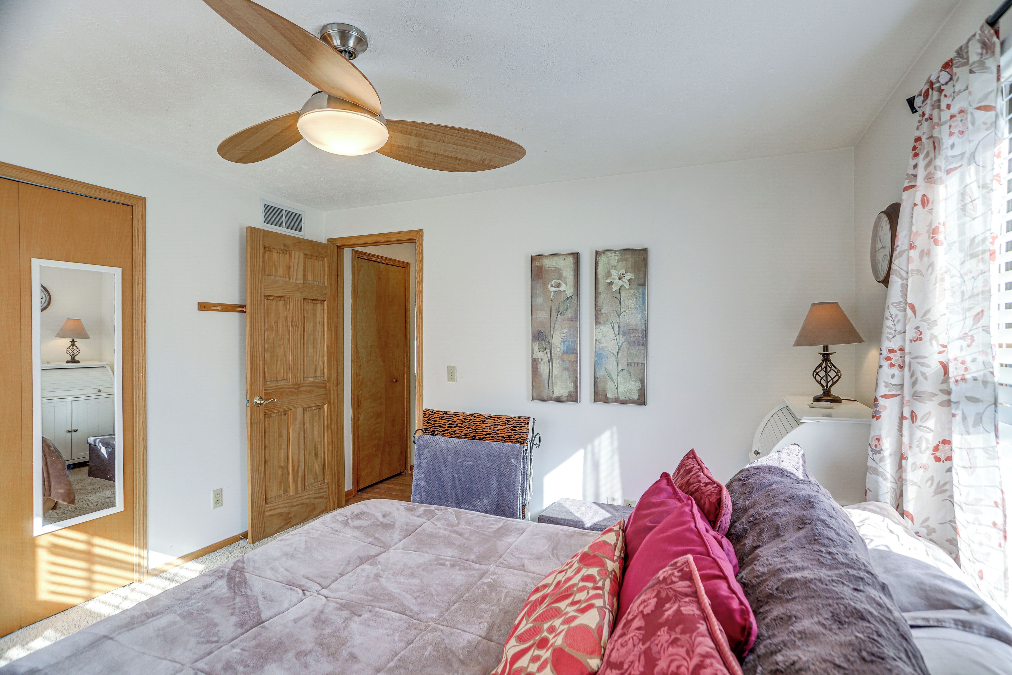 A bedroom with a bed, a ceiling fan, a door, a mirror, and a wall clock.