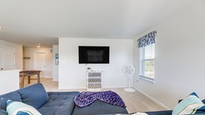 A modern living room with a grey sectional sofa, a flat-screen TV mounted on the wall, a small side table with a decorative item, and a window with blinds. - Game Room + Patio: Spacious Ocean View Home (Ocean View)