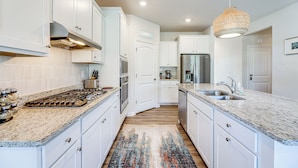 A kitchen with white cabinets, granite countertops, a stainless steel refrigerator, and a woven pendant light. - Game Room + Patio: Spacious Ocean View Home (Ocean View)