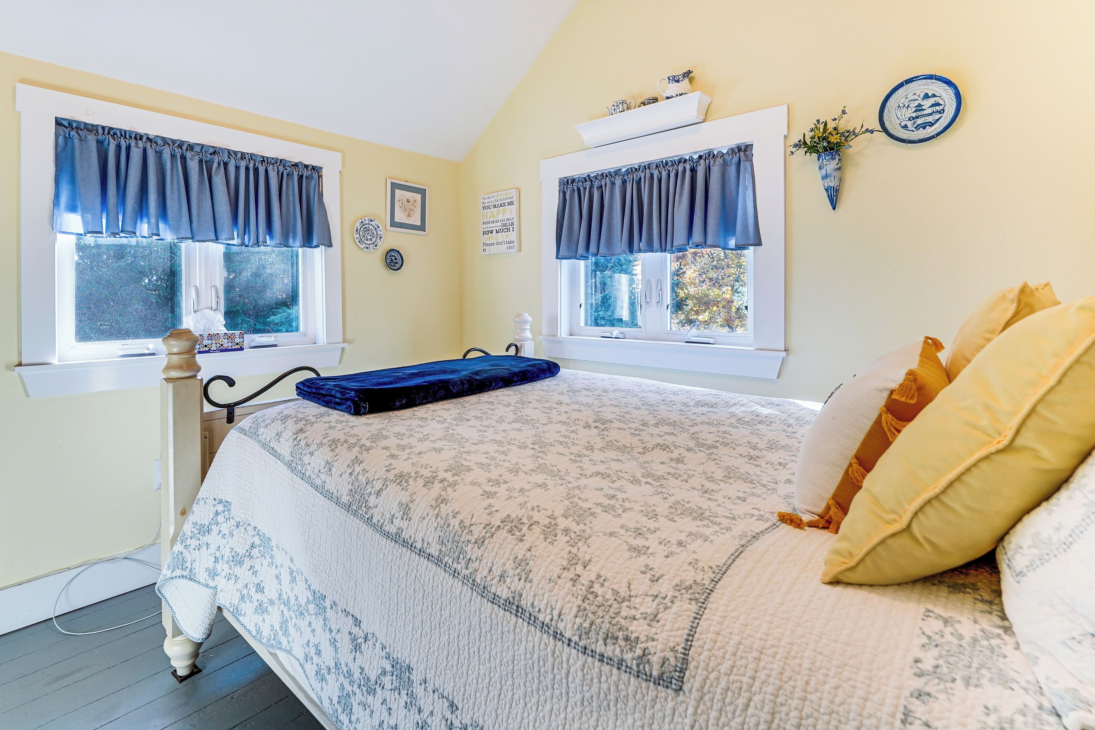 A bedroom with a bed, two windows with curtains, a bedside table, and a wall-mounted shelf with decorative items.