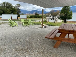 Eine Kiesterrasse mit einem Holztisch und grünen Stühlen, einem überdachten Bereich sowie Blick auf Berge und einen See.