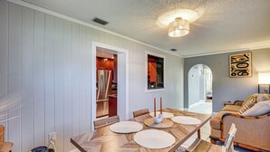 Uma sala de jantar com uma mesa de madeira posta para quatro pessoas, um sofá e uma cozinha visível através de uma porta aberta.