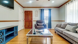 Una sala de estar con una mesa de centro de vidrio, un sofá de cuero, un sillón de cuero, un televisor montado en la pared y un ventilador de techo.