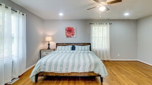 Un dormitorio con una cama de madera, un ventilador de techo, una mesita de noche con lámpara y un ventanal con cortinas translúcidas.