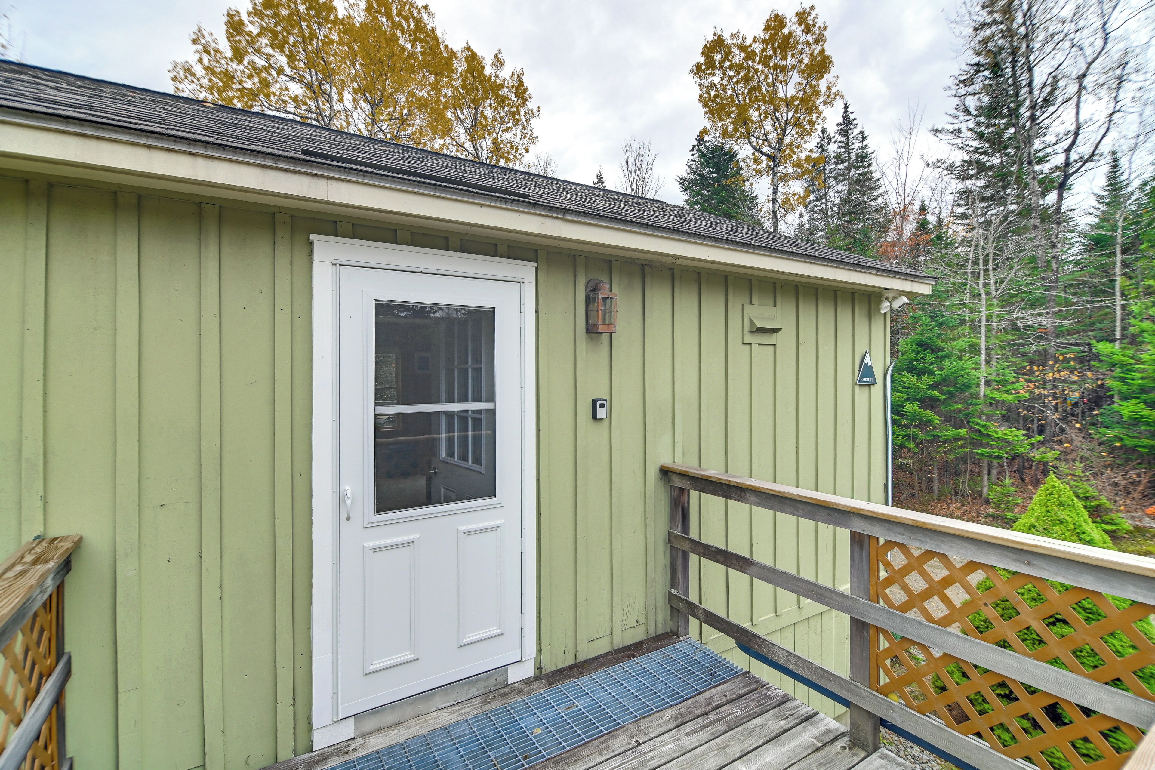 A small, single-story building with a green exterior, a white door, and a wooden deck with a lattice railing.