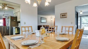 Un coin repas avec une table en bois dressée pour quatre personnes, entourée de chaises. On aperçoit un réfrigérateur et une lampe de table dans le fond.