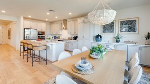 A modern kitchen with white cabinetry, a central island, and a dining area with a wooden table and chairs. - Elegant & Spacious Retreat w/ Yard in Woodland! (Woodland)