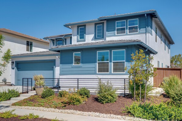 A two-story house with a garage, a landscaped front yard with various shrubs and a small tree, and a clear blue sky. - Elegant & Spacious Retreat w/ Yard in Woodland! (Woodland)